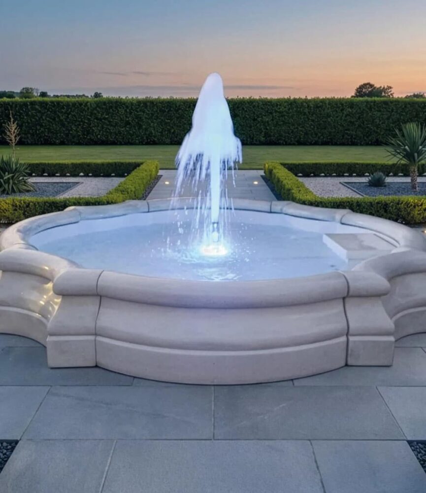 Fontana circolare da giardino in pietra con bacino sagomato, illuminazione interna e getto d'acqua centrale.
